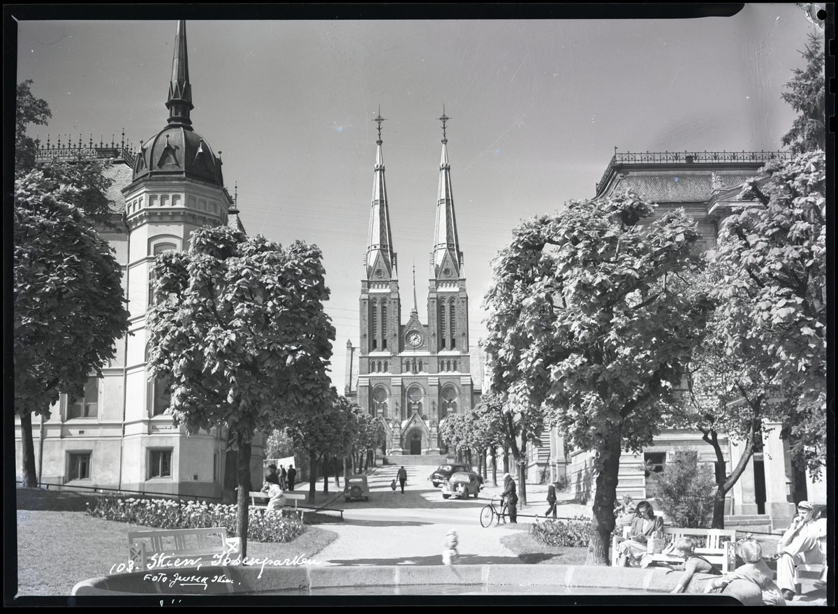 Skien kirke tatt fra Ibsenparken i sentrum av byen Til høyre (bak trærne) kan man se deler av Festiviteten til høyre og Skien Sparebank til venstre.