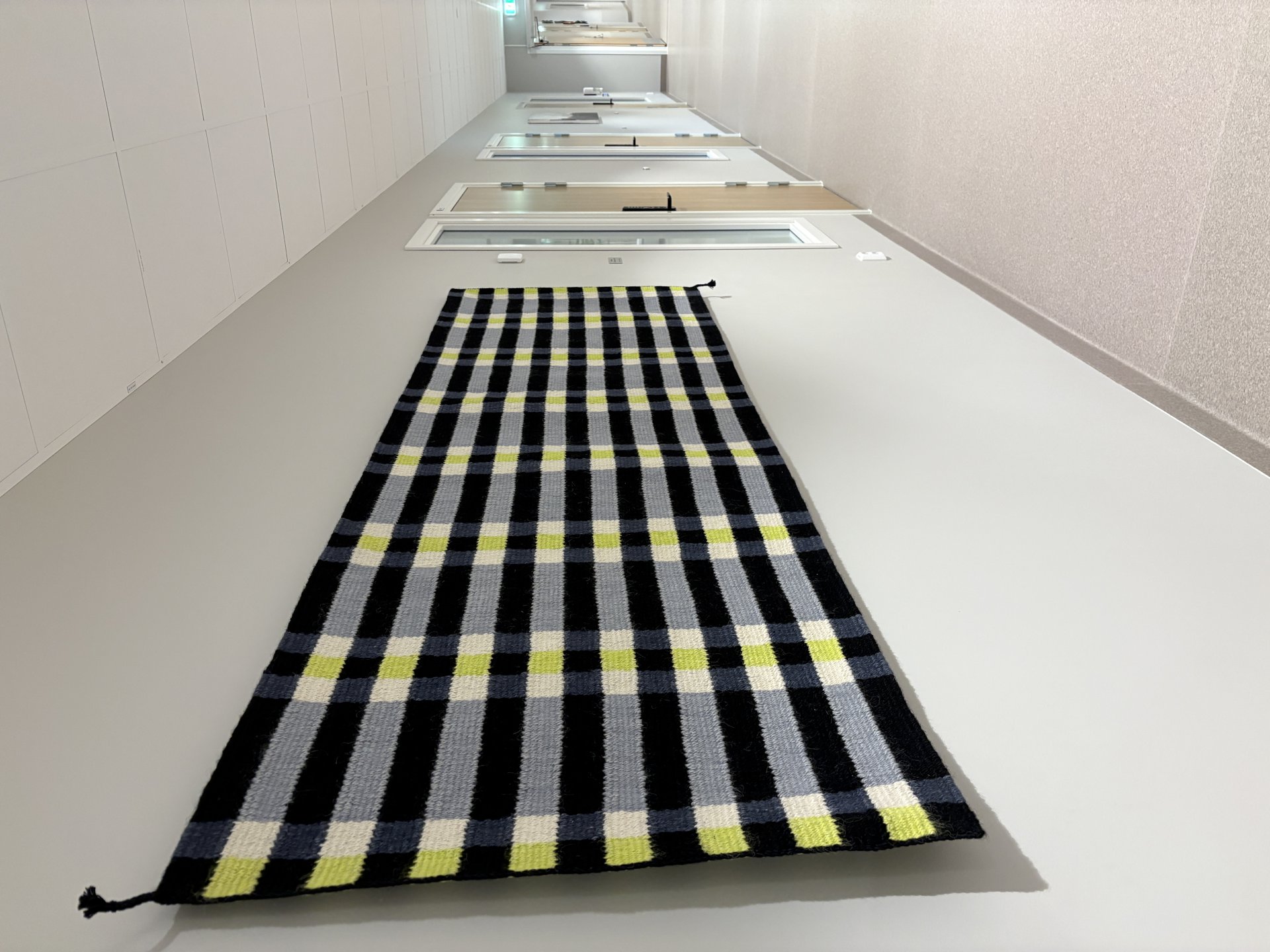 Tekstilteppe i gult, sort, hvitt og grått som henger på vegg i en korridor.